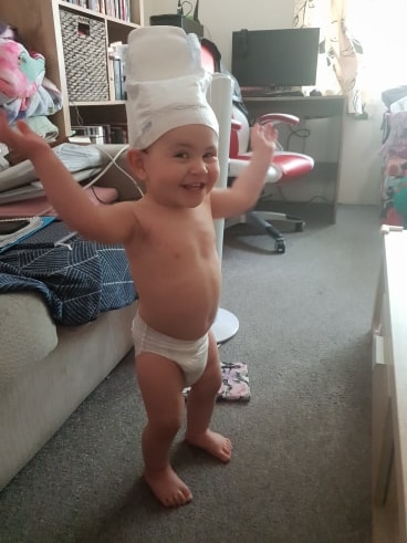 A happy baby stands in a lounge room, wearing a disposable nappy on her head.