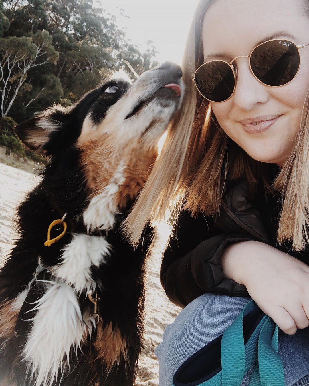 A selfie of a blonde woman wearing sunglasses, with a black tan and white kelpie licking her cheek