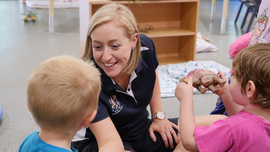 A blonde childcare worker plays with two children, sitting on the ground.