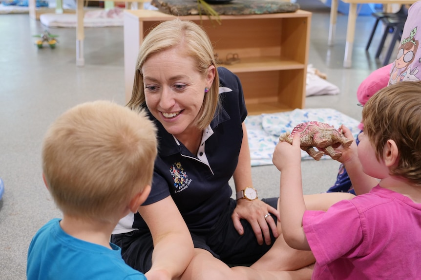 A blonde childcare worker plays with two children, sitting on the ground.