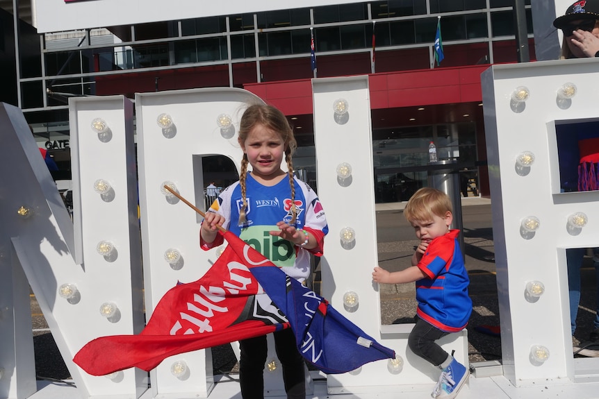 A young girl with a Newcastle knights flag stands outside a stadium.