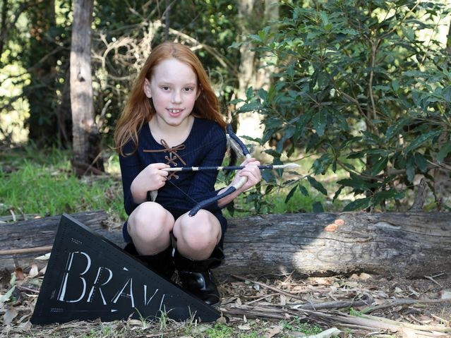 Jesse smiles, holding a fake bow and arrow with a sign that says 'brave'.