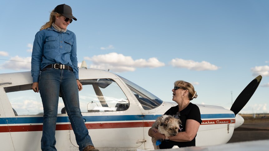 A young blonde woman stands on the wing of a small plane and smiles at a woman standing on the ground, holding a fluffy dog
