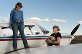 A young blonde woman stands on the wing of a small plane and smiles at a woman standing on the ground, holding a fluffy dog