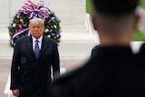 Donald Trump stands infront of a wreath as rain falls on his suit and a soldier salutes in the foreground