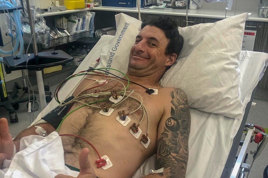 A man lies in a hospital bed with monitoring wires on his chest, he is grimacing with his thumbs up and looks uncomfortable