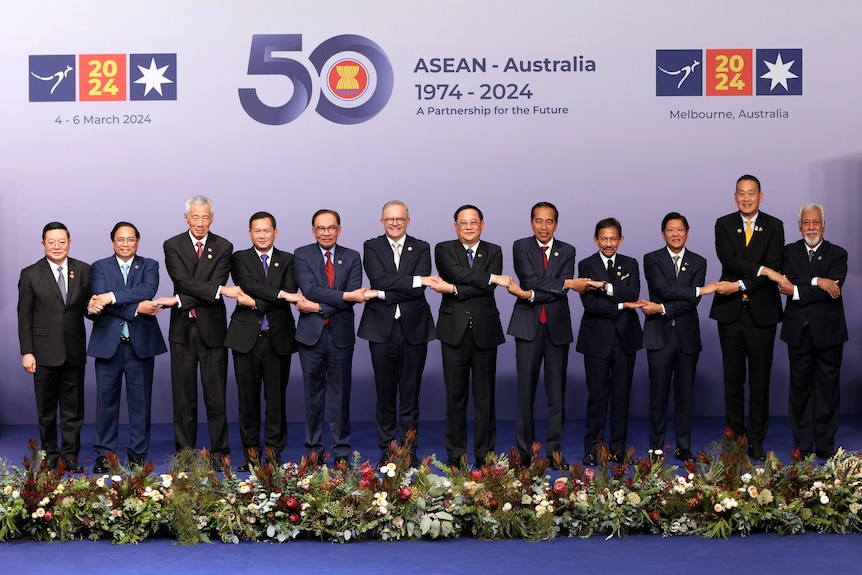 Twelve male leaders from ASEAN nations wear dark suits and hold hands as they stand in a line and pose for a photo.