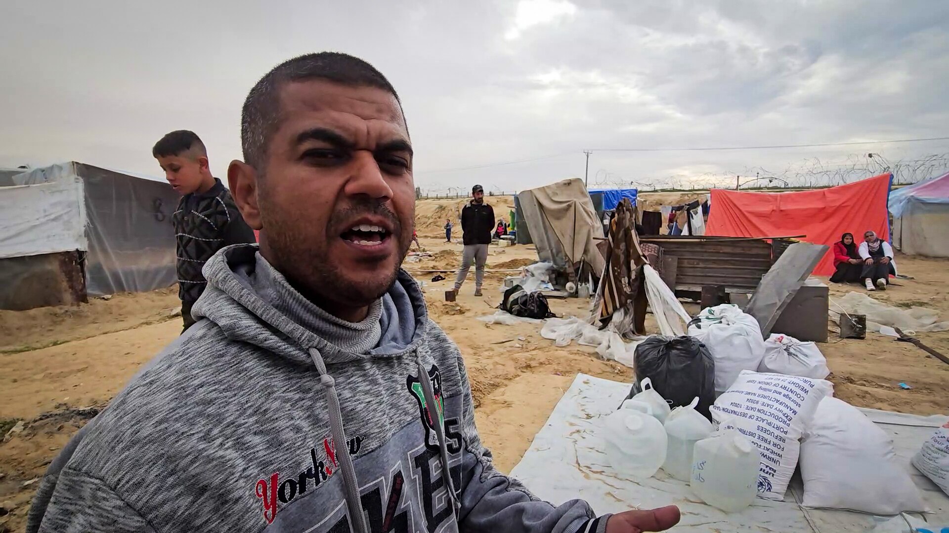 A man looks down the barrel of the camera, speaking animatedly in front of a tarp with scattered belongings