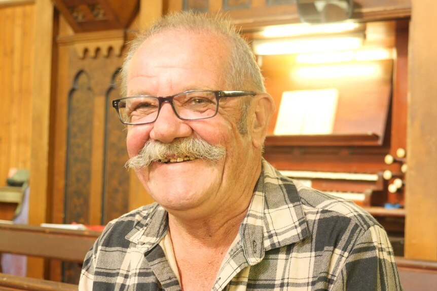 A man with a grey mustache smiles in front of a wooden organ