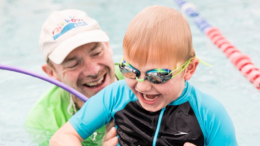 A man in a white cap and child with goggles swim in a pool.
