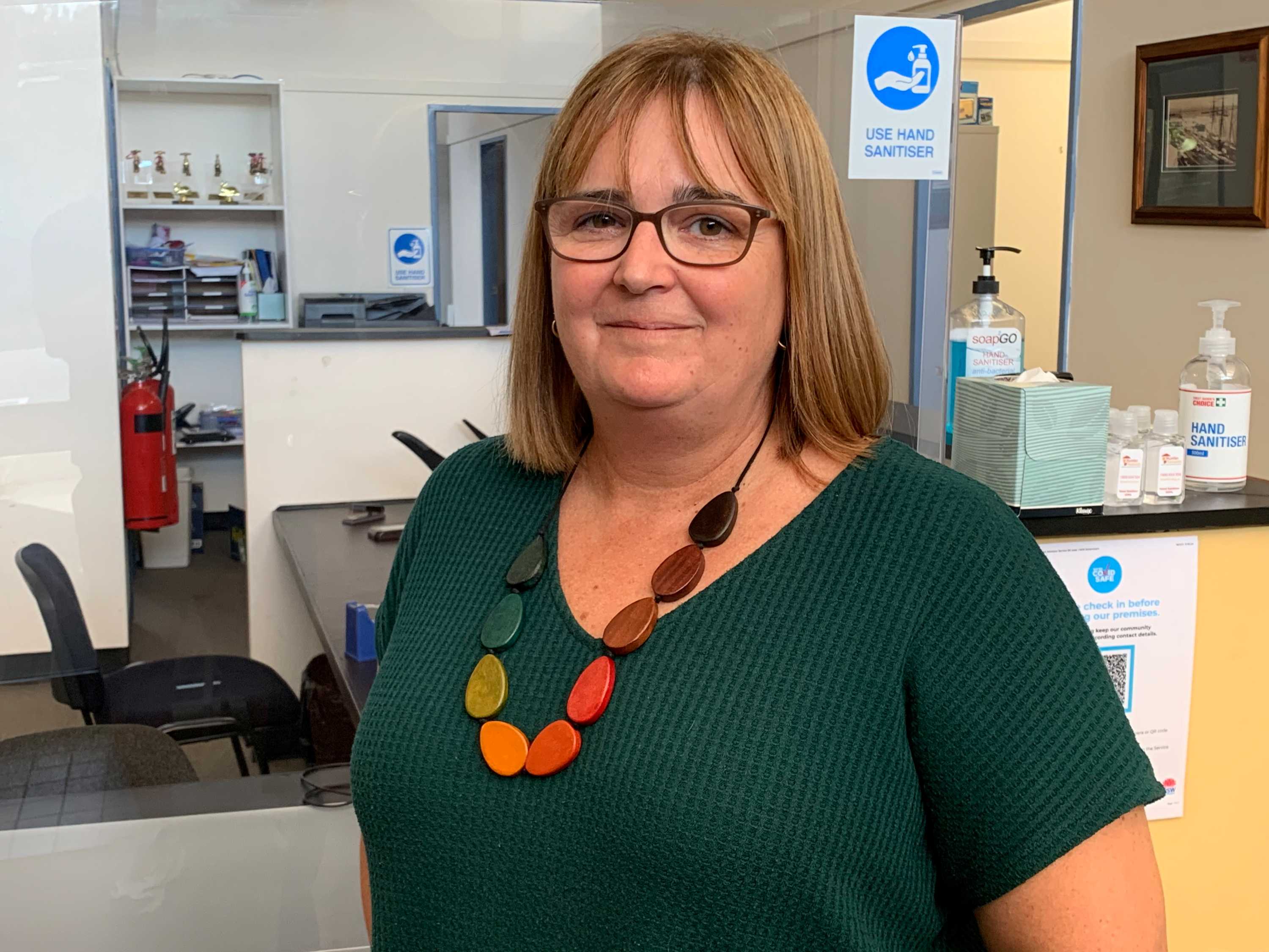 A woman wearing brown glasses, a green top and colourful necklace looks at the camera.