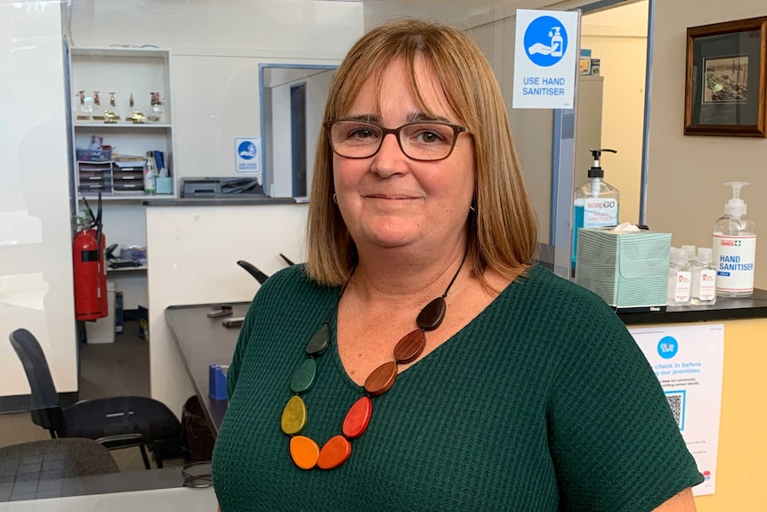 A woman wearing brown glasses, a green top and colourful necklace looks at the camera.