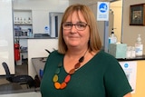 A woman wearing brown glasses, a green top and colourful necklace looks at the camera.