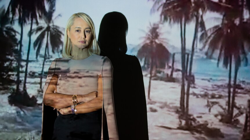 Rebekah stands in front of a projection on a wall of a beach being destroyed by a tidal wave