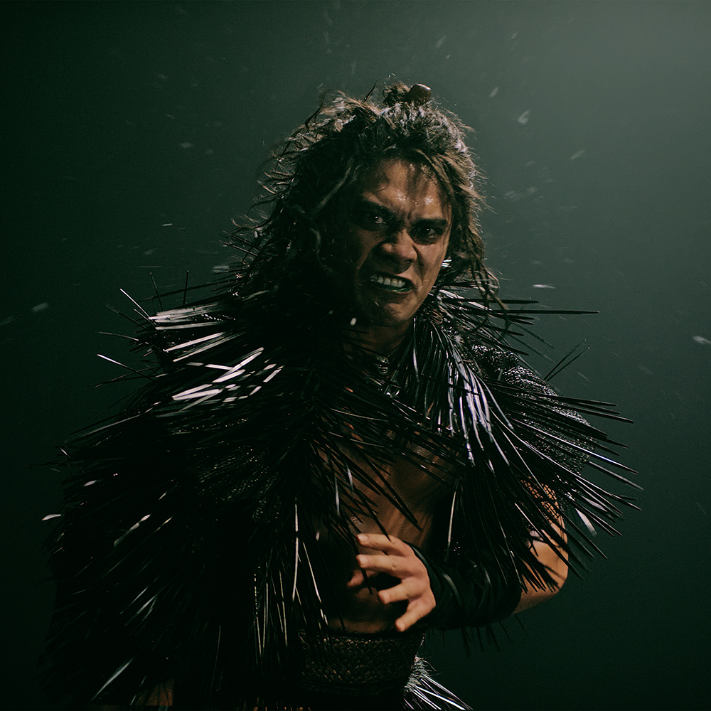 A man with long dark hair with fierce expression wears black spikey garment and stands ready for a physical confrontation.