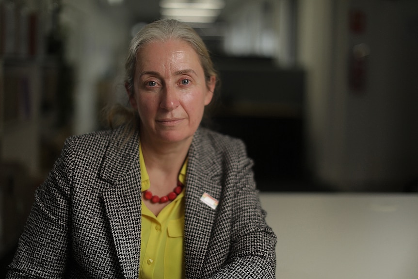 A woman in business attire, looks into the camera with a neutral expression. Behind her is a darkened office.