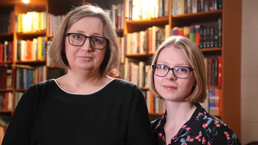 Two women with blonde hair and wearing black glasses stand in front of a bookshelf