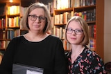 Two women with blonde hair and wearing black glasses stand in front of a bookshelf