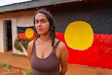 A young woman in a brown singlet stands in front of an Aboriginal flag mural. 