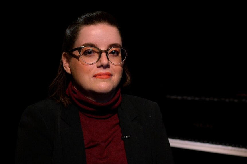 A woman with glasses in a red turtle-neck and black jacket in a dark room with piano behind