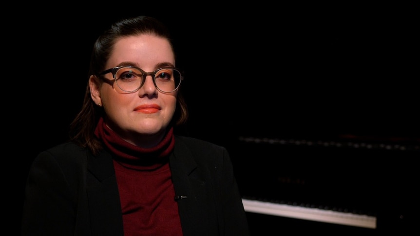 A woman with glasses in a red turtle-neck and black jacket in a dark room with piano behind