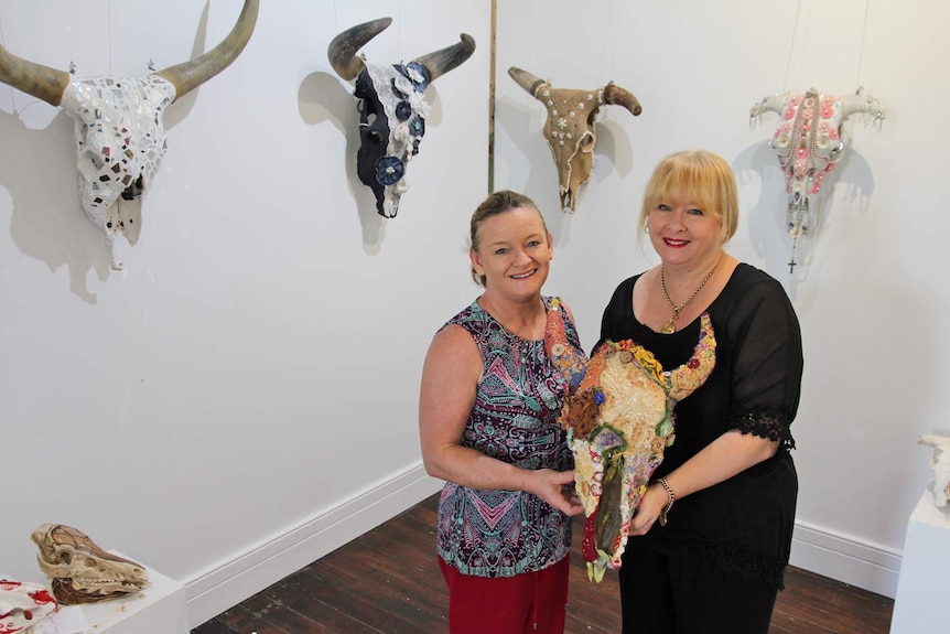 Two women hold a decorated skull and stand in a gallery space with many decorated skulls