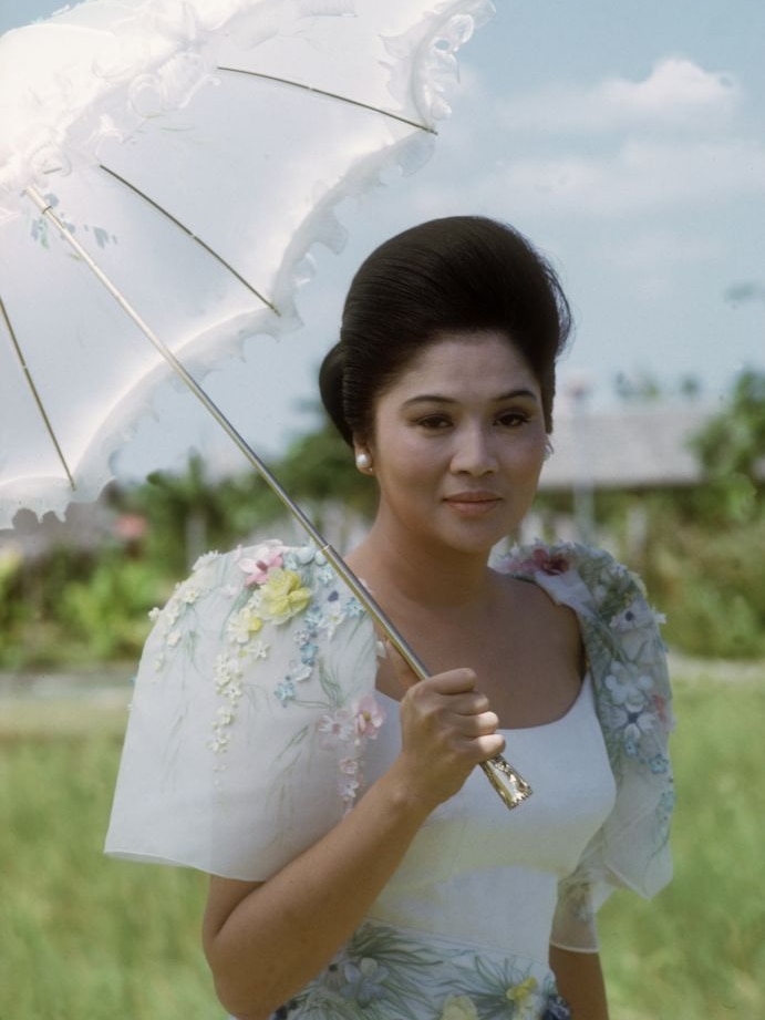 You view a portrait of a young Imelda Marcos in a bucolic scene, wearing a Terno and holding a white, semi-transparent umbrella.