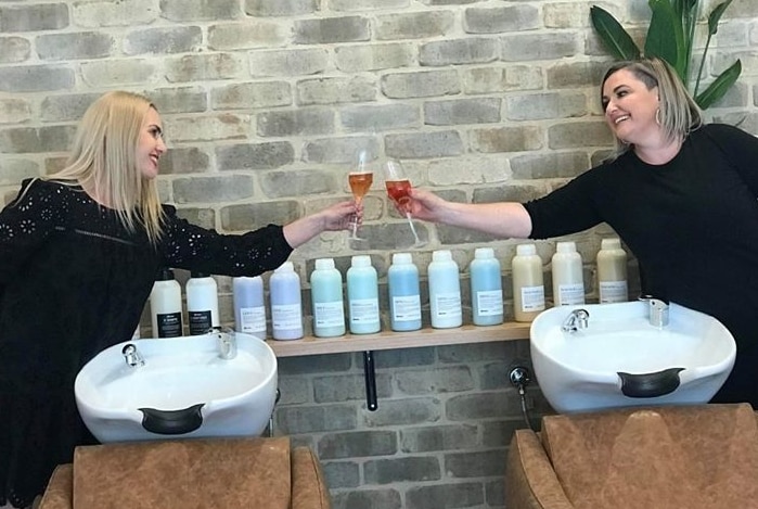 Two ladies, social distancing, looking at each other and reaching out to clink their drink glasses.