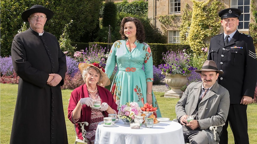 A photograph of characters from TV series Father Brown, in 50s costume, standing and seated for tea in an English garden
