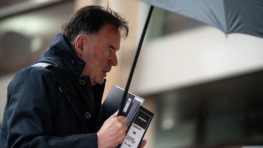 Man walking into court holding documents and covering his face with an umbrella.