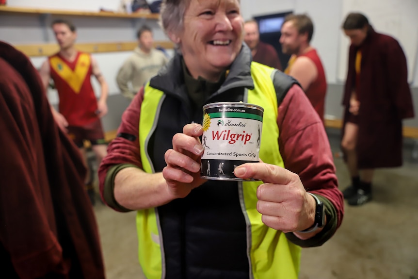 An older woman holds a tin of sports grip product in a football club changerooms with football players behind her