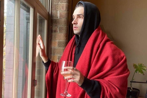 Louis Hanson looks longingly out the window, while covered in a blanket and holding wine. 
