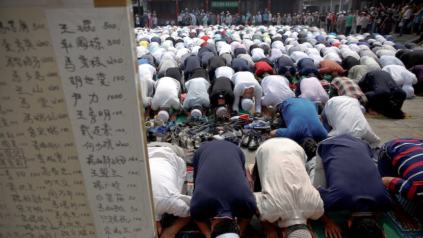 A large group of men prostrate themselves in prayer behind a board of Chinese writing.