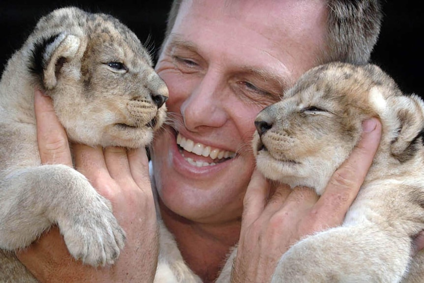 A photo of Tim Husband smiling while holding two lion cubs.