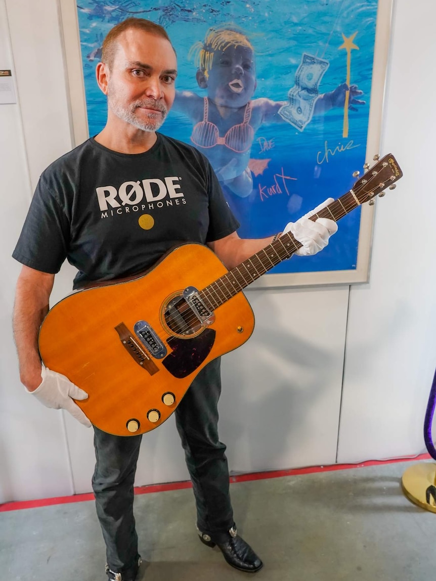 Peter Freedman holds the guitar while standing in front of a poster featuring the cover of Nevermind