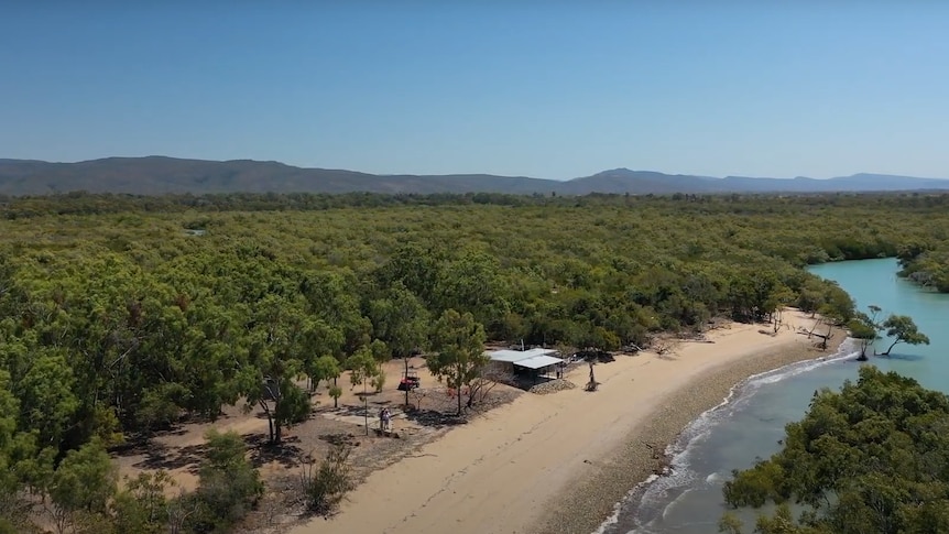 Ariel picture of a house sitting on the edge of a beach with trees and mountains behind. 