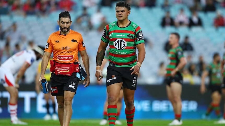 South Sydney NRL star Latrell Mitchell looks serious as he walks off the ground with a trainer during an NRL game. 