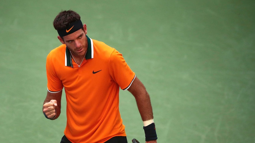 A male tennis player in an orange shirt clenches his fist and looks off to the side