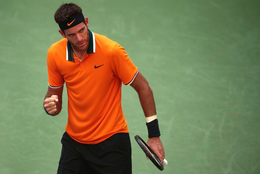 A male tennis player in an orange shirt clenches his fist and looks off to the side