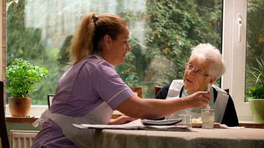 A woman in an apron assisting an elderly woman with her meal at a table in front of a window