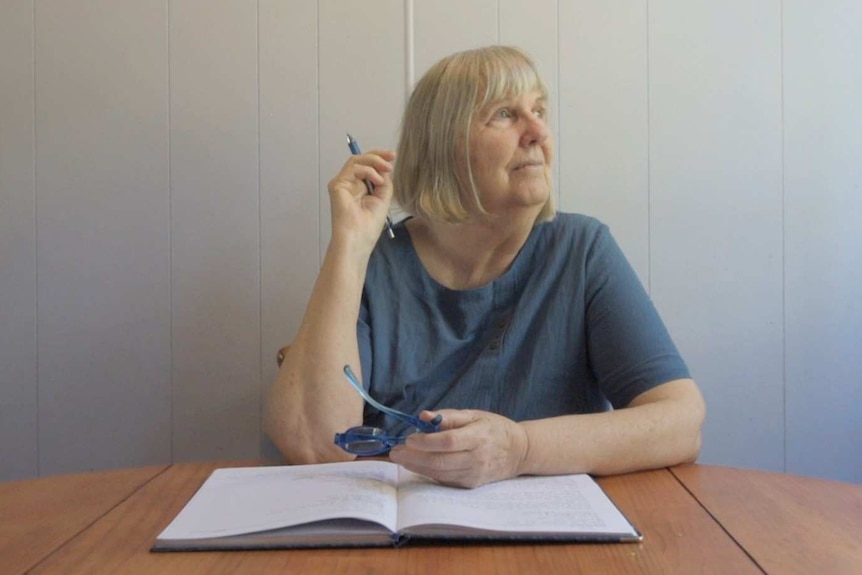 Melita Luck sits at her wooden dining table in a blue shirt looking out to the left holding a pen with a notebook on the table