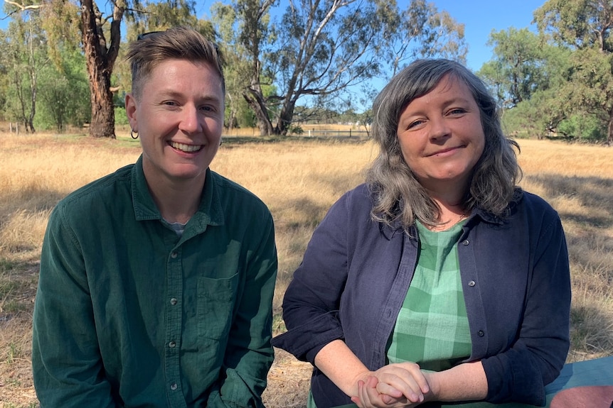 Two people sitting, smiling at camera with a farm property background. 
