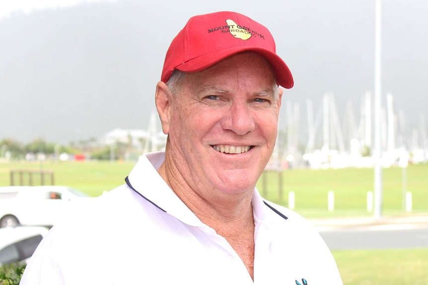 A man wearing a bright cap and a light-colored polo shirt sands smiling in front of what appears to be a marina.