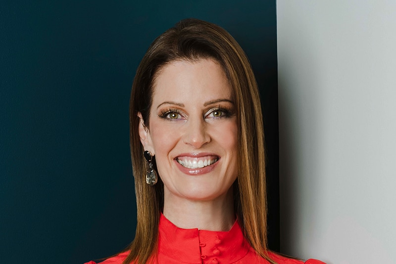 Cate Bakos smiles, wearing a red top indoors.