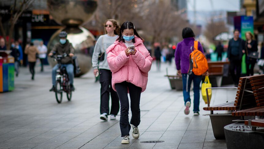 A woman wearing a big pink puffy jacket walks through Adelaide's Rundle Mall while wearing a face mask and using a mobile phone