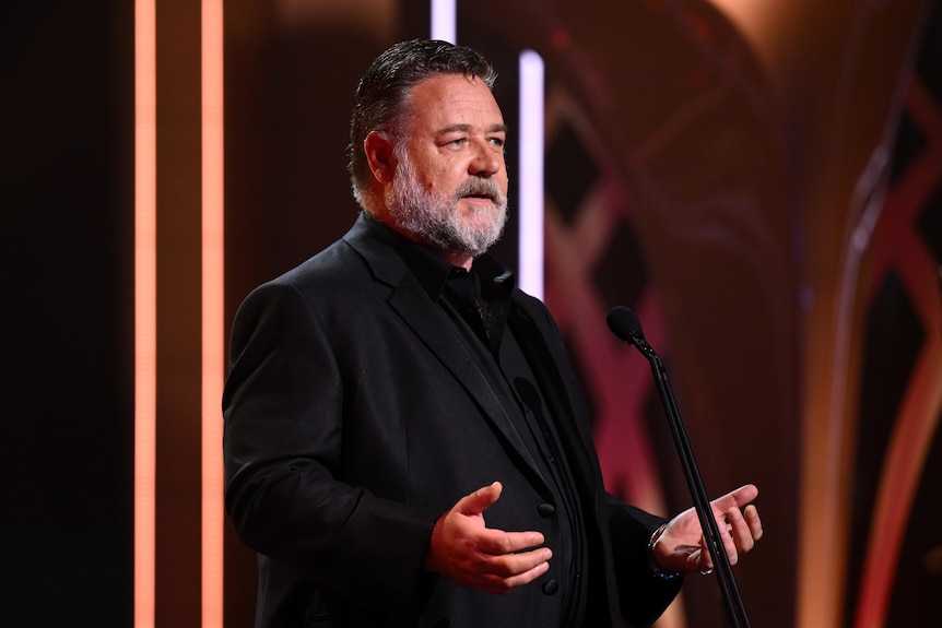 Russell Crowe, a middle-aged white man with greying hair and beard, wears a black suit and speaks on a stage.