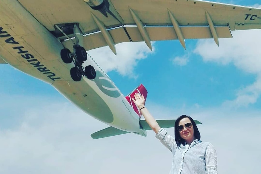 A dark-haired woman reaches up and appears to almost touch a plane flying low overhead