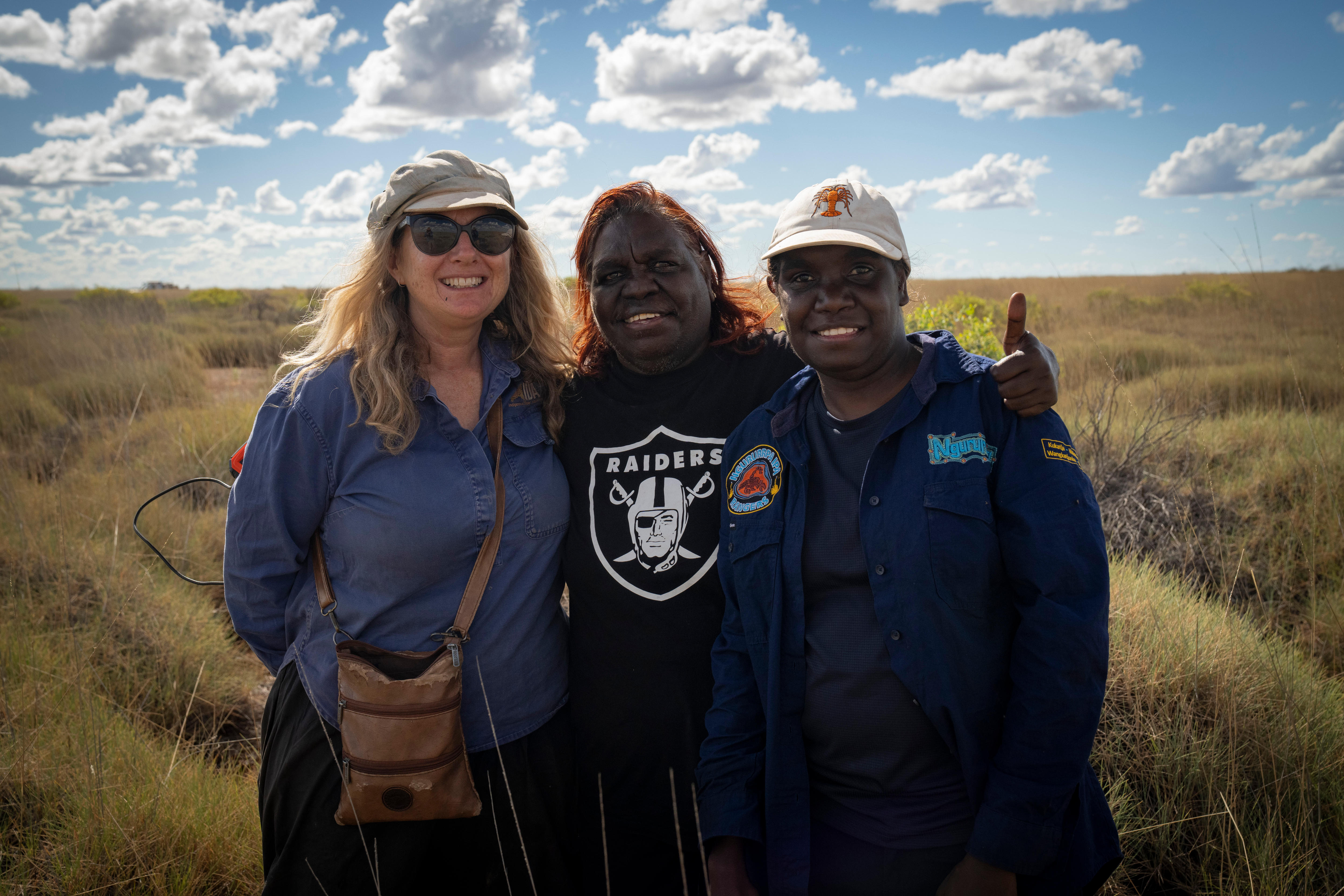 A white woman with sunglasses, a middle-aged Aboriginal woman and a younger Aboriginal woman stand together by spinifex.