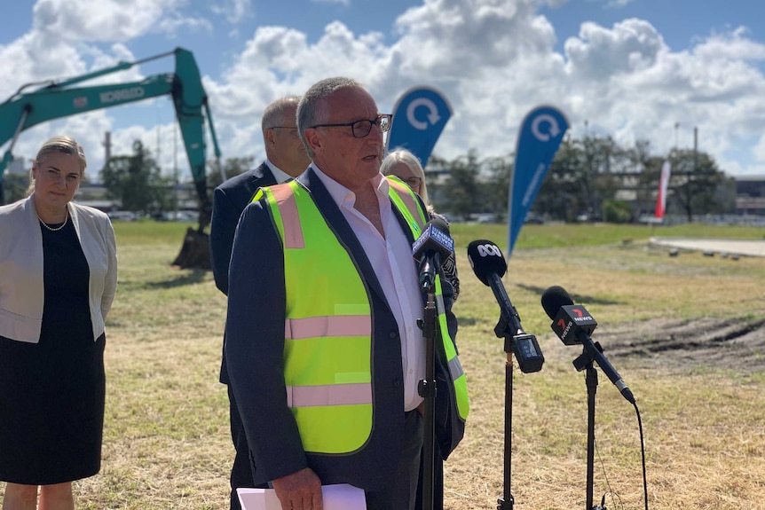 A man wearing a hi-vis vest stands in front of several microphones in a paddock.