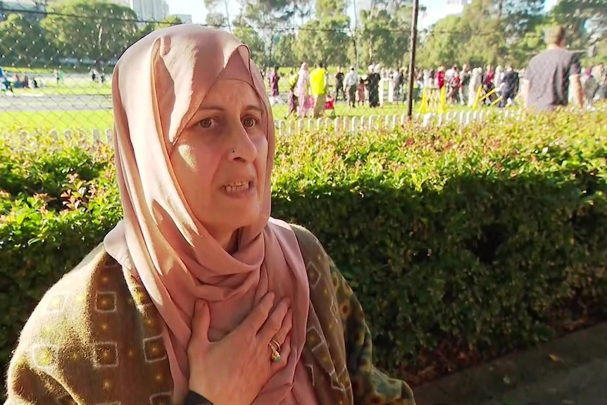 A woman wearing a hijab puts her hand to her chest as she speaks, with a row of hedges behind her and a crowd in the distance.